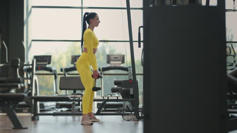 Young-Hispanic-brunette-woman-squats-holding-a-dumbbell-in-the-gym-in-a-yellow-tracksuit